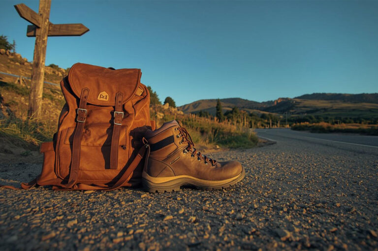 Brown leather hiking boot and a matching vintage backpack resting on a gravel roadside near a wooden signpost in the mountains, symbolizing hiking and adventure travel.