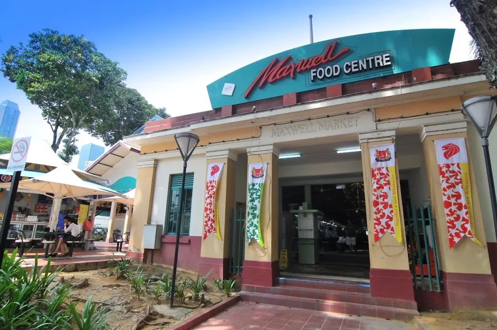 The image features the prominent entrance of the Maxwell Food Centre, displaying a large teal and red sign above a cream and red building facade labeled "Maxwell Market." Colorful banners hang from the pillars flanking the entrance, while outdoor seating under umbrellas and lush green foliage are visible to the left under a bright blue sky.