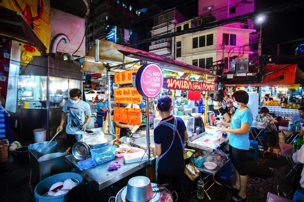 A vibrant night scene at a bustling outdoor food stall in Yaowarat, where vendors and customers wearing face masks interact amidst steaming pots and brightly lit signs. In the background of streets, multi-story city buildings are illuminated by colorful pink and white street lights under the dark sky.