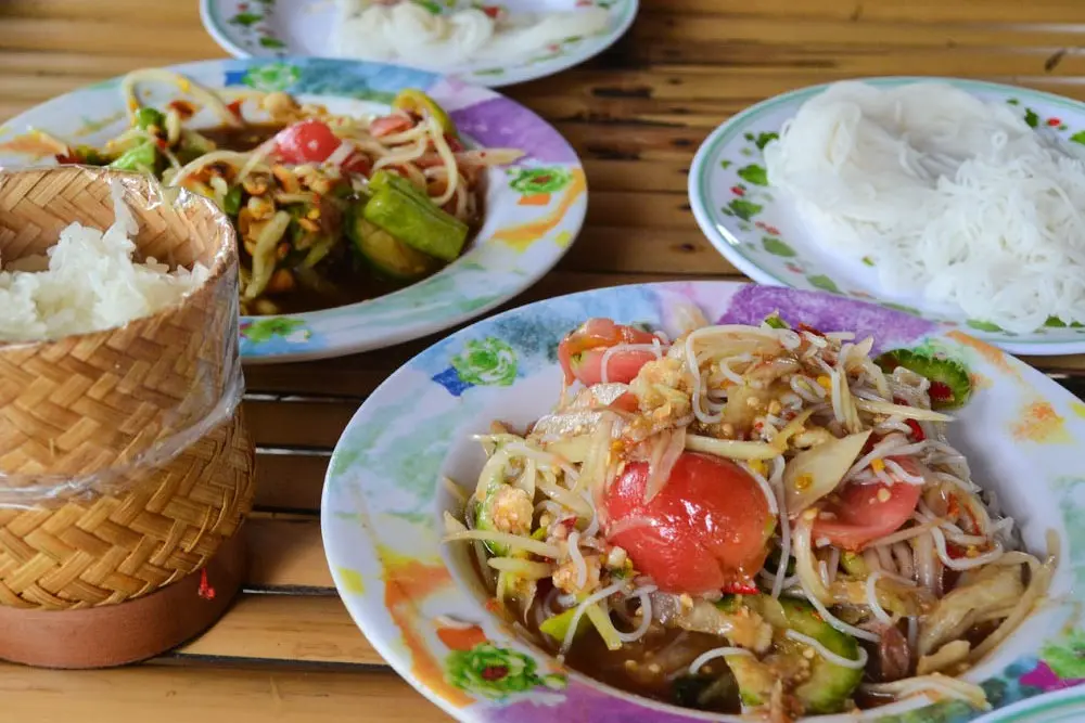 A vibrant spread of Thai Isaan cuisine features plates of spicy green papaya salad mixed with rice noodles, fresh tomatoes, and long beans on a bamboo table. Beside the main dishes sits a traditional small woven bamboo basket filled with sticky rice, accompanied by additional side plates of plain white rice noodles.