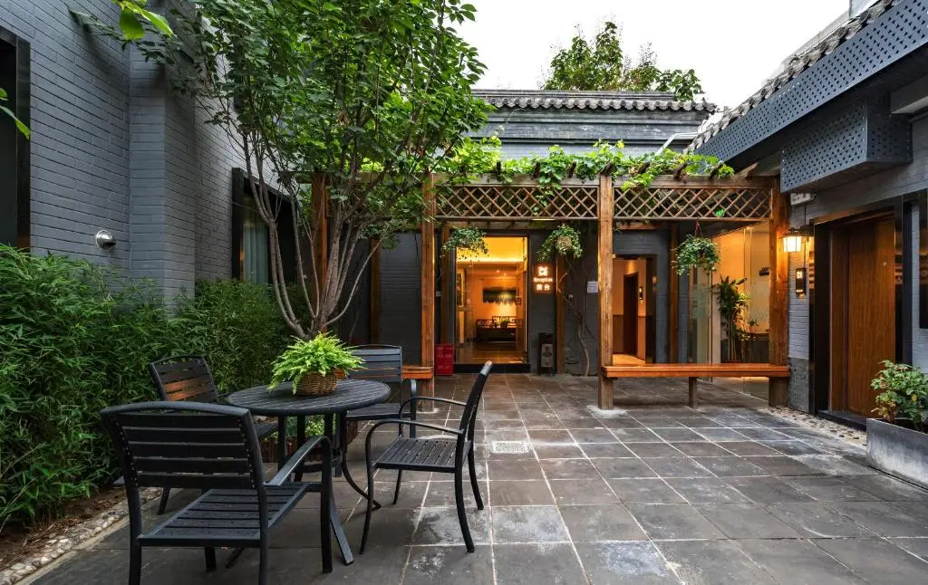 Alt. Text for 312195769.jpg: An outdoor courtyard paved with dark gray tiles features a black patio set with a round table and matching chairs under the shade of a leafy green tree. In the background, a wooden pergola covered in climbing vines sits in front of building entrances with warm interior lighting.