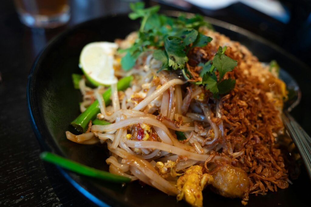 A steaming plate of Pad Thai is topped with fresh cilantro, a lime wedge, and a generous portion of crispy fried shallots. The dish features rice noodles mixed with egg, green onions, and savory proteins in a dark ceramic bowl.