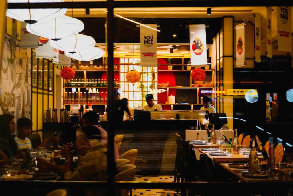 The image shows the warm, brightly lit interior of a Japanese restaurant called "You Mee," featuring paper lanterns and chefs working behind a sleek service counter. Customers are seated at tables in the foreground, captured through a window that reflects the surrounding urban environment.