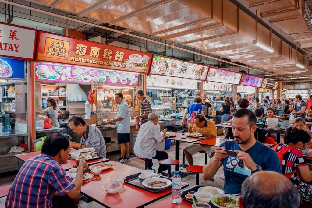 This vibrant image captures a bustling scene inside a traditional Singaporean hawker center, where diners sit at red tables enjoying various local dishes like Hainanese Chicken Rice. Brightly lit food stalls with colorful signage line the background, as vendors prepare meals and people of various backgrounds engage in the lively atmosphere of communal dining.