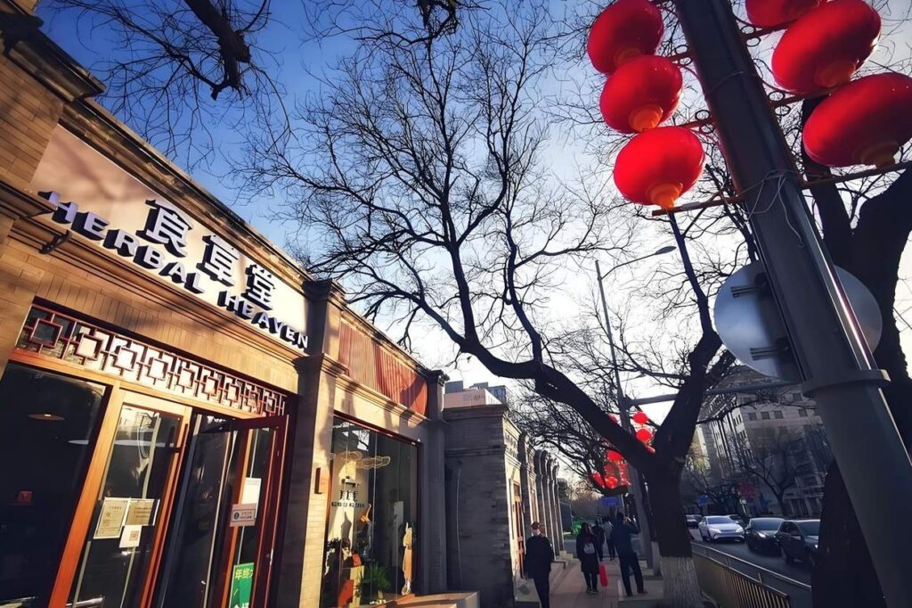 The image captures a street scene in Beijing featuring a store named "Herbal Heaven" set against traditional grey-brick architecture and bare winter trees. Vibrant red lanterns hang from a pole in the foreground, contrasting with the blue sky and the soft late-afternoon light hitting the sidewalk.