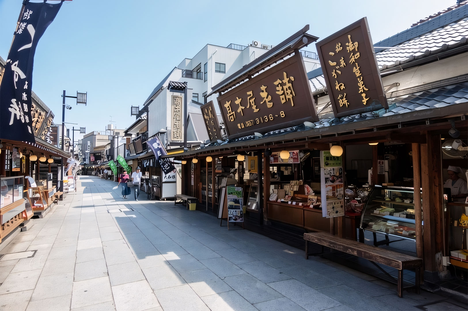 Traditional, quiet Japanese street (shitamachi) with historic wooden merchant shops and large signage, likely leading to a temple or popular tourist site on a sunny day.