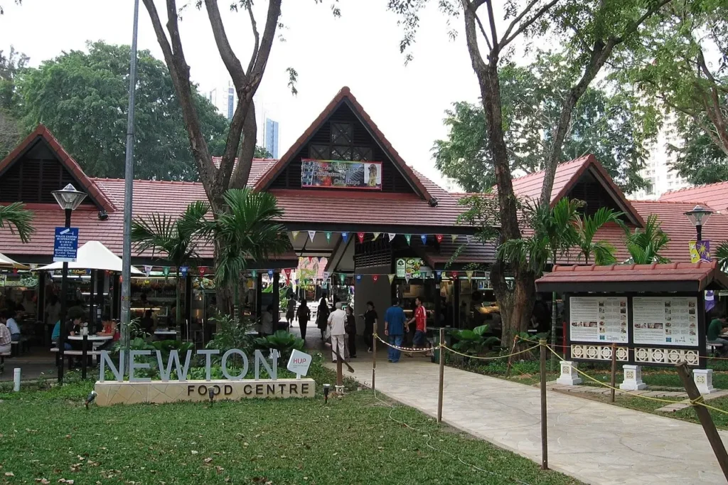 The image captures the entrance of the open-air Newton Food Centre, featuring a large grey sign spelling out its name on a grassy lawn. A paved pathway guides visitors toward the food stalls, which are housed under traditional red-tiled roofs and shaded by large, mature trees.