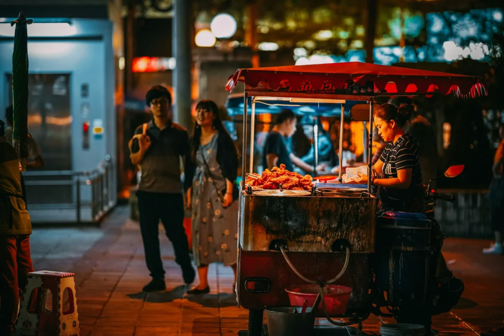 In the image, a woman stands under the red canopy of a street food cart at night, preparing fried chicken for customers. In the softly blurred background, pedestrians walk along the illuminated city sidewalk under warm streetlights.
