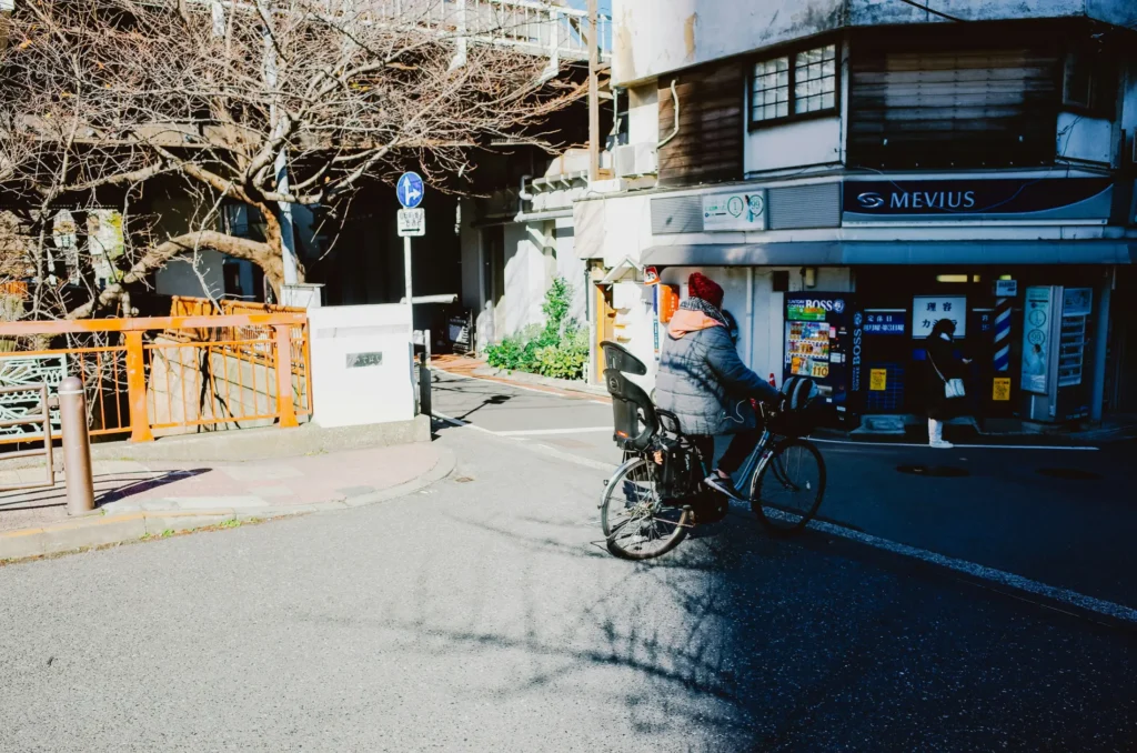 In the image, a person wearing a grey winter jacket and a red knit beanie rides a bicycle equipped with a child seat down an urban street. The background features bare winter trees, an elevated structure, and a storefront lined with Japanese vending machines under a "MEVIUS" sign.