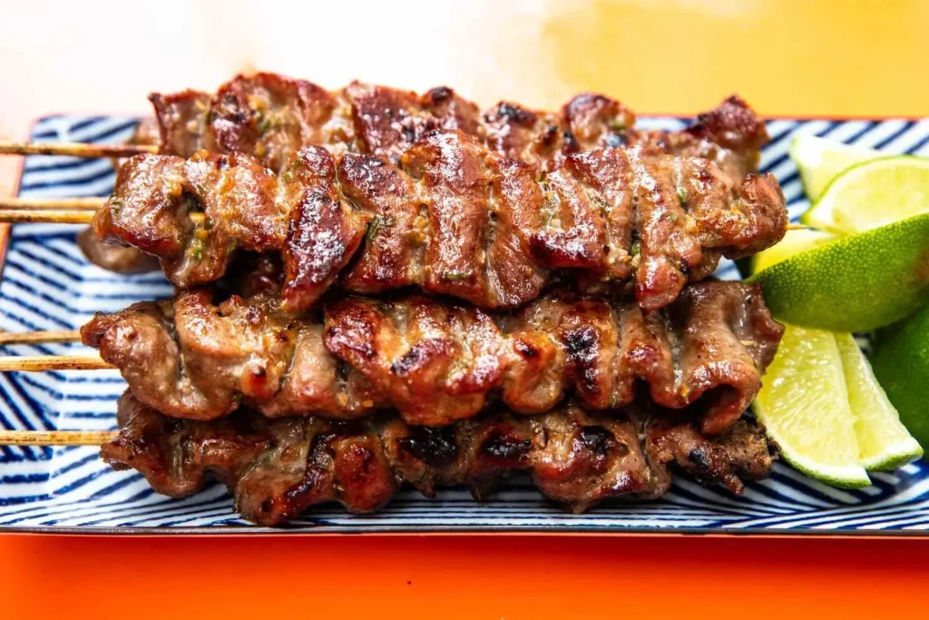 A close-up shot displays a stack of freshly grilled, glistening Thai pork skewers arranged on a blue-and-white patterned plate. The savory skewers are served alongside bright lime wedges against a vibrant orange background.