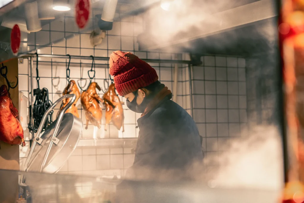A worker wearing a red beanie and black face mask looks downward while working behind a glass window obscured by thick cooking steam. In the background against a white tiled wall, several golden-brown roasted ducks hang from metal hooks.