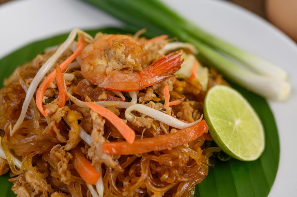 A vibrant plate of Pad Thai is served on a green banana leaf, featuring stir-fried glass noodles topped with a large succulent shrimp and thin strips of carrot. The dish is accompanied by a fresh lime wedge and whole green onions, all presented on a clean white plate.