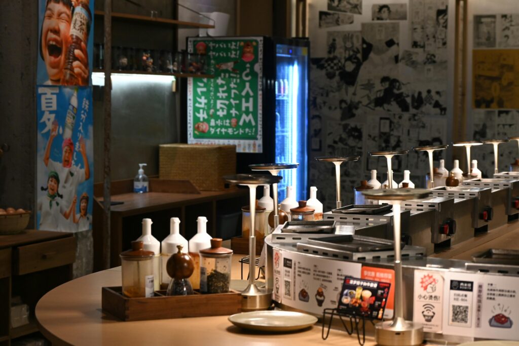 The image shows the warm, brightly lit interior of a Japanese restaurant called "You Mee," featuring paper lanterns and chefs working behind a sleek service counter. Customers are seated at tables in the foreground, captured through a window that reflects the surrounding urban environment.