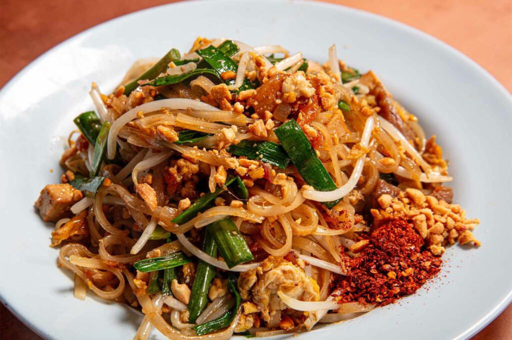 Close-up of a generous serving of Pad Thai noodles on a white plate, topped with bean sprouts, chives, peanuts, and a pile of red chili flakes for seasoning.