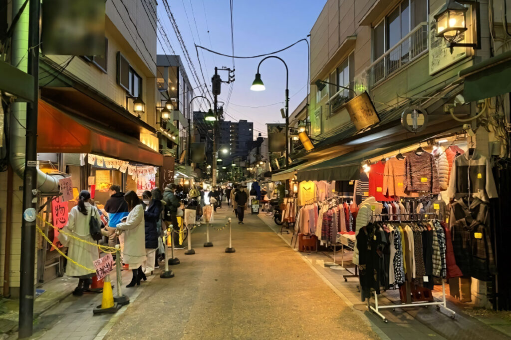 Narrow, bustling Japanese shopping alleyway at dusk, lined with small shops, clothing racks, and warm lantern lighting.