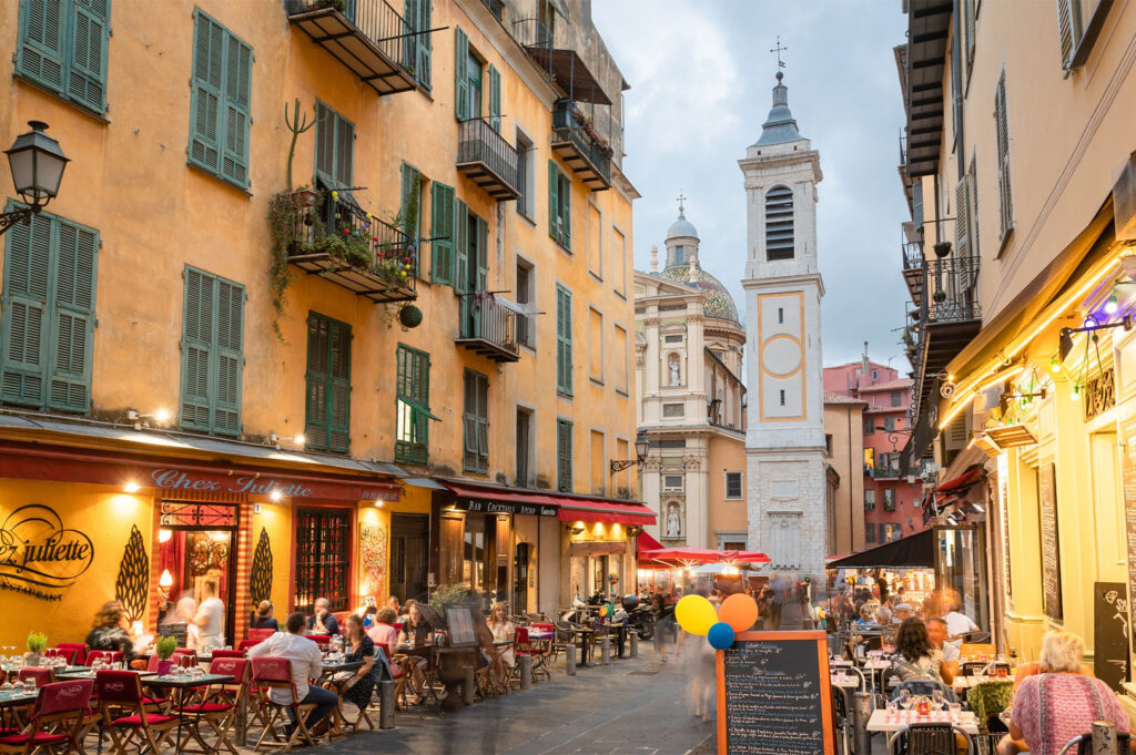 Lively, narrow street in Nice Old Town (Vieux Nice) with yellow buildings, green shutters, outdoor patio dining, and the Baroque clock tower of Chapelle de la Miséricorde.