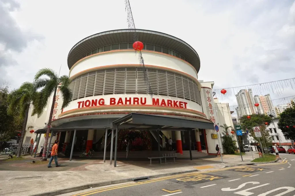 The image features the distinctive, curved art deco facade of the Tiong Bahru Market under a cloudy sky. The entrance is marked by large red lettering, flanked by palm trees, red Chinese lanterns, and a street with a "Pick Up" zone in the foreground.