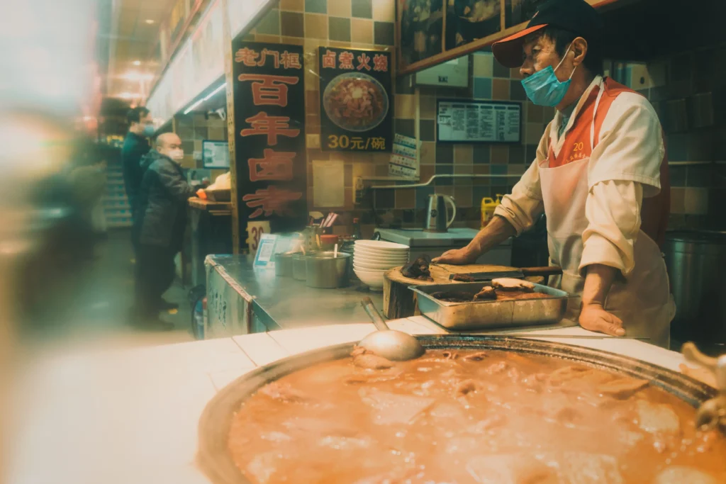 An older male worker wearing a blue face mask, cap, and apron stands behind a food counter next to a large pot of simmering stew. In the background of the warm-toned eatery, Chinese signage and posters are visible on the tiled wall alongside other masked patrons.
