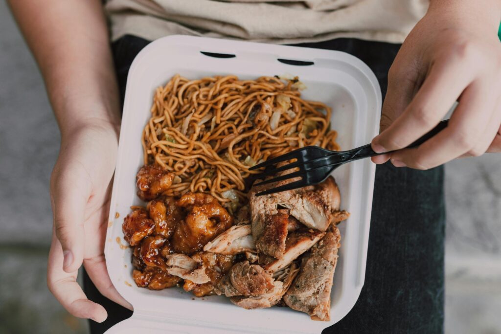 This image shows a close-up overhead view of a person holding a white takeaway container filled with a hearty meal of noodles and two types of meat. Using a black plastic fork, they are about to eat a portion of the sliced roasted meat, which sits alongside saucy orange chicken and a bed of stir-fried noodles.