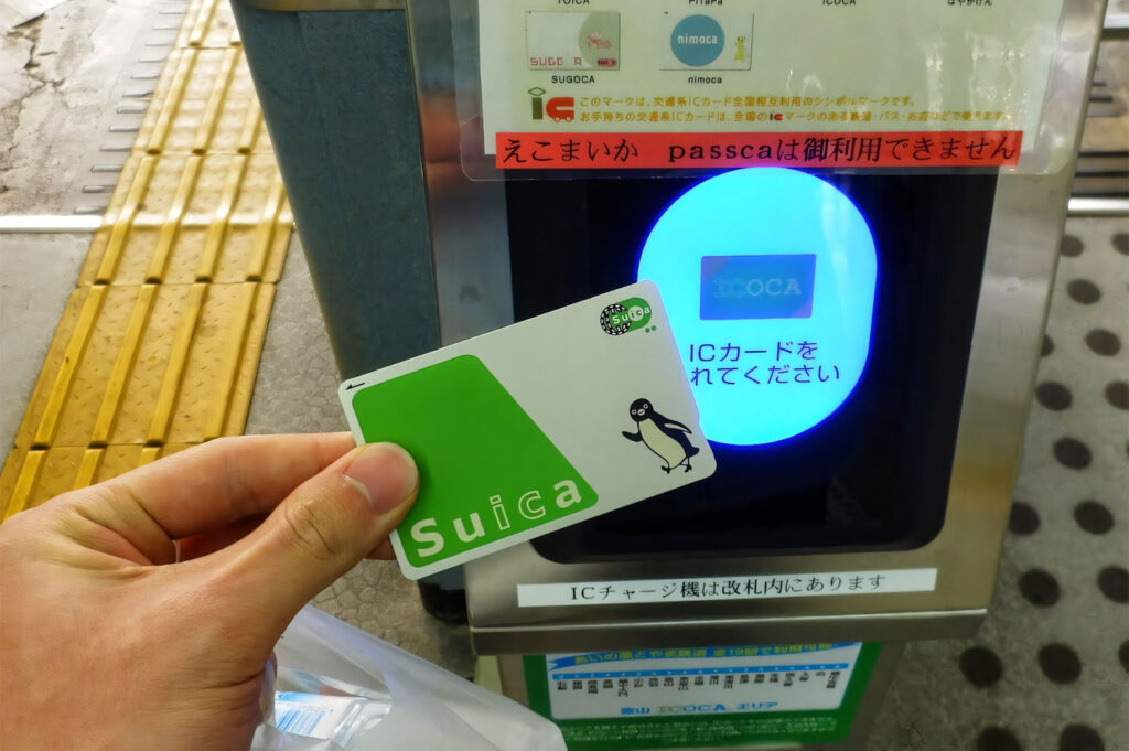 A person holding a green Suica IC card up to a glowing blue transit reader at a Japanese train or subway station ticket gate.