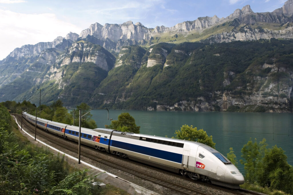 A French TGV high-speed train traveling along the shore of a large lake, set against a dramatic backdrop of steep, rocky mountains (likely in the French Alps/Swiss border region).