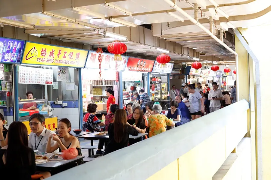 The image shows a bustling open-air food center lined with vibrant food stalls featuring Chinese signage and red lanterns. Numerous patrons are seated at long tables enjoying their meals, while others stand and walk along the busy corridor.