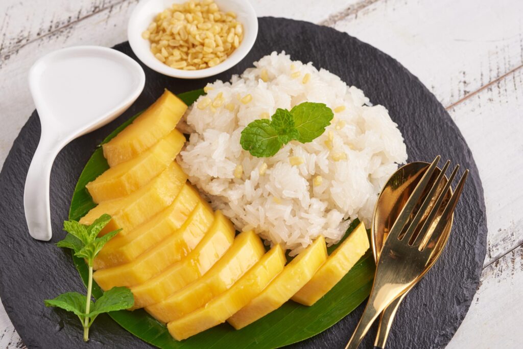 A beautiful serving of mango sticky rice is displayed on a dark slate plate, featuring sliced ripe mango and sweet rice garnished with mint leaves. Small side dishes of creamy coconut milk and toasted mung beans complete the traditional Thai dessert presentation.