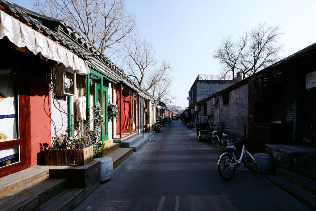 The first image features the ornate facade of a historic building in Beijing, marked by a large black sign with gold calligraphy draped in red fabric. The second image captures the imposing Drum Tower of Xi'an, a multi-tiered wooden structure resting on a massive stone base under a soft, late-afternoon sky. The final image shows a narrow, quiet hutong alley lined with traditional grey-brick buildings, colorful storefronts, and parked bicycles under a clear blue sky.