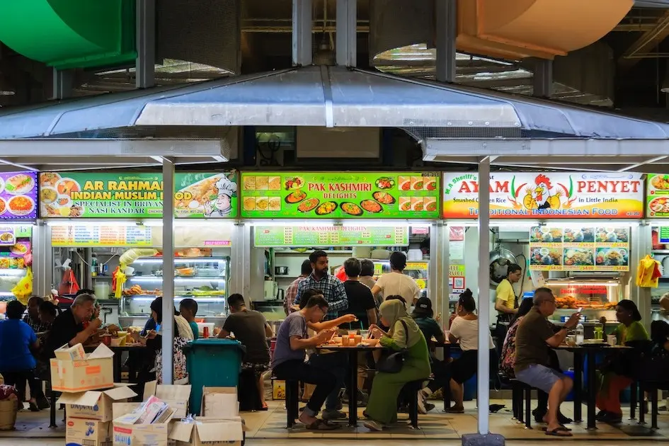 The image captures a bustling hawker center with brightly lit food stalls serving Indian Muslim, Kashmiri, and Indonesian cuisines. Numerous patrons are seated at tables in the foreground, dining and socializing in a lively communal atmosphere.