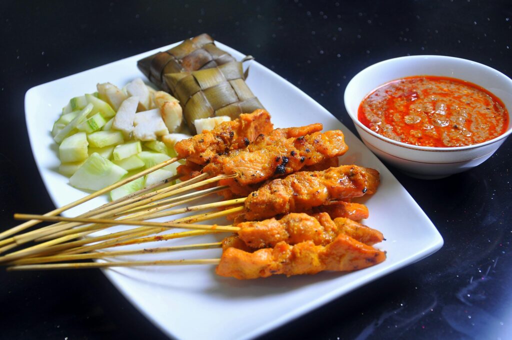 A plate of grilled chicken satay skewers is served with traditional sides of compressed rice cakes (ketupat) and fresh cucumber chunks. A bowl of rich, spicy peanut sauce sits next to the platter, ready for dipping.