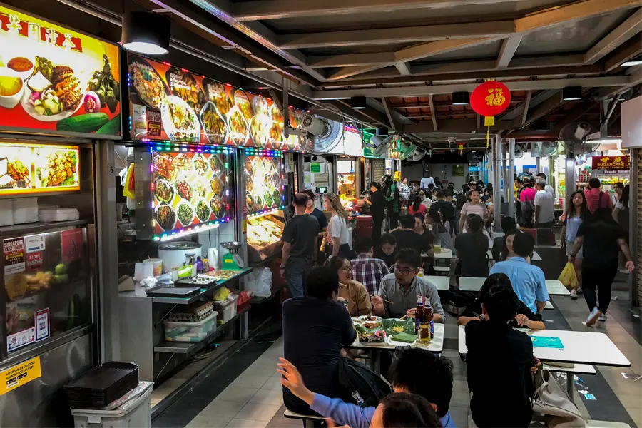 The image captures a bustling, vibrant indoor hawker center filled with numerous patrons seated at white tables enjoying their meals. A row of food stalls lines the left side, featuring brightly illuminated menus with colorful photos of various dishes beneath a covered ceiling with a red hanging lantern.