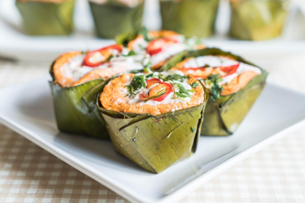 This image shows several portions of Khanom Krok, a Thai steamed fish curry mousse, served in traditional handcrafted banana leaf cups. Each cup is garnished with a drizzle of coconut cream, sliced red chilies, and fresh herbs to highlight its vibrant orange color.