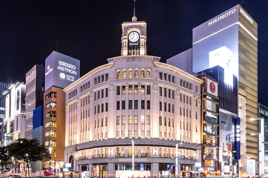Nighttime view of the iconic Wako building with its clock tower in Ginza, Tokyo, surrounded by brightly lit modern luxury retail buildings, including SEIKO and MIKIMOTO.