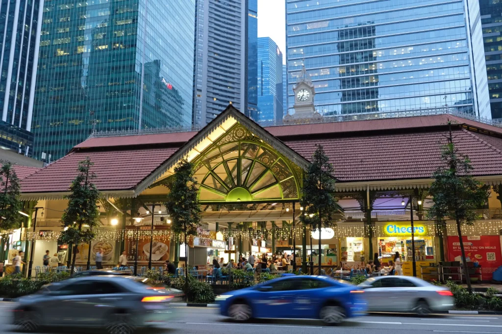 This image showcases the historic Lau Pa Sat hawker center nestled in the heart of Singapore's financial district. The vibrant food market features a traditional tiled roof and intricate green archways, standing in sharp contrast to the towering modern glass skyscrapers and blurred passing traffic in the foreground.