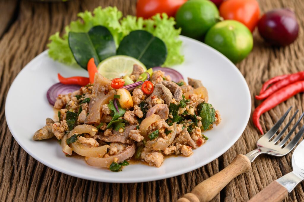 This vibrant plate features a spicy minced pork salad, known as Larb, garnished with fresh mint, lime slices, and red onion. The dish is served on a rustic wooden table alongside fresh ingredients like chilies, tomatoes, and whole limes.