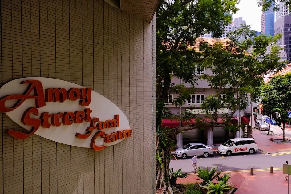 An oval sign for the "Amoy Street Food Centre" is mounted on a tiled wall in the foreground on the left. In the background, a street view shows parked cars, a pedestrian, and lush green trees framing traditional buildings and distant city skyscrapers.
