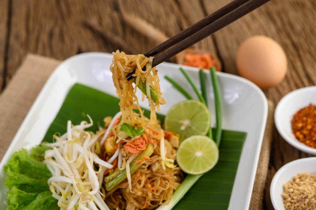 This image features a vibrant plate of Pad Thai served on a banana leaf with fresh bean sprouts, lime wedges, and green onions. Chopsticks lift a bundle of the stir-fried noodles, while side bowls of crushed peanuts and chili flakes sit ready for seasoning on the wooden table.