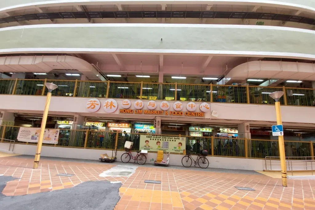 The image shows the exterior entrance of the multi-story "Hong Lim Market & Food Centre" building with its sign written in both Chinese characters and English. Bicycles and delivery carts are parked along the tiled open-air walkway in front of the fenced lower level stalls.