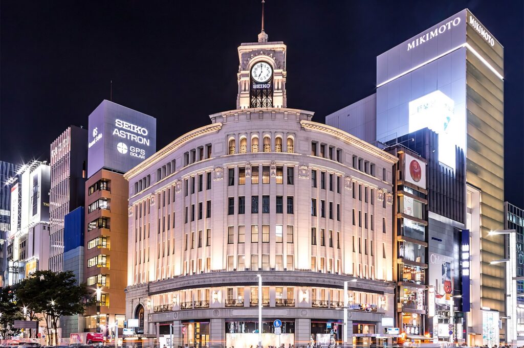 Nighttime view of the iconic Wako building with its clock tower in Ginza, Tokyo, surrounded by brightly lit modern luxury retail buildings, including SEIKO and MIKIMOTO.