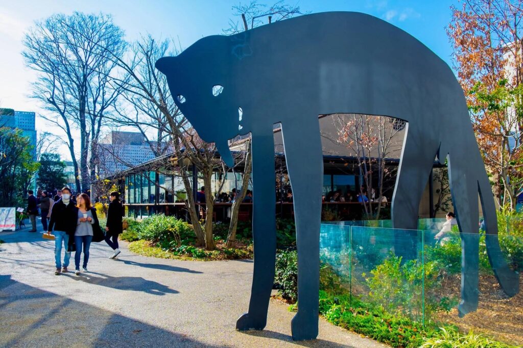 A large, stylized black metal sculpture of a cat stands prominently in a sunlit outdoor plaza, towering over a paved walkway and nearby greenery. Pedestrians stroll through the open area in the background, which features a modern glass-walled building nestled among bare winter trees under a clear blue sky.