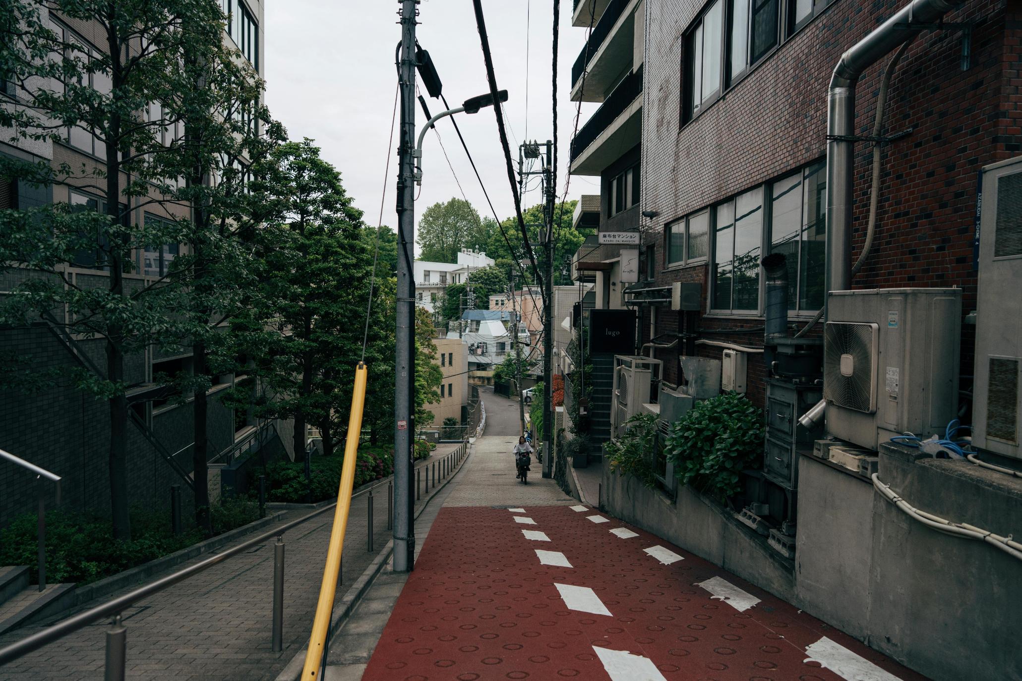 This narrow, sloping street in Japan features a distinctive red-textured pavement marked with white rectangles and a cyclist riding in the distance. Densely packed urban elements frame the scene, including brick and concrete buildings, tangled utility lines, and lush green trees lining the hillside.