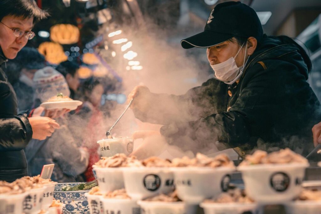 This photograph captures a busy street food vendor, wearing a dark jacket, a baseball cap, and a surgical mask, serving a customer from a steaming hot pot or dish.