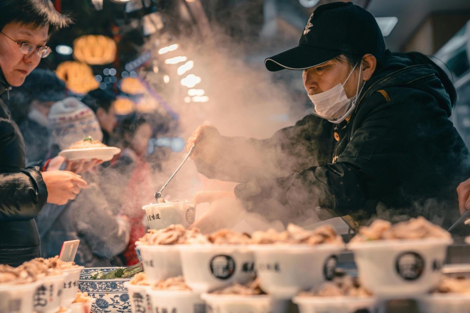 This photograph captures a busy street food vendor, wearing a dark jacket, a baseball cap, and a surgical mask, serving a customer from a steaming hot pot or dish.