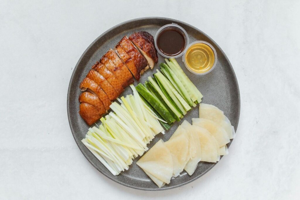 This overhead image displays a plate featuring the components of a Peking Duck meal, arranged neatly on a dark gray, textured platter against a light background.