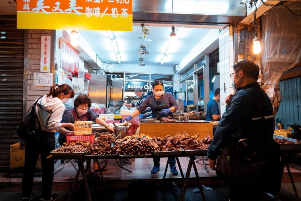 This image captures a brightly lit street food stall at night, where three vendors, all wearing masks, are serving customers from an abundant display of skewers and cooked meats on a table in the foreground.