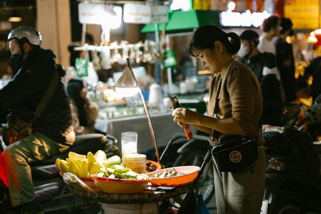 This image features a street vendor, dressed in a brown knitted top, standing next to her cart and actively using her mobile phone under the glow of a single hanging lamp at night.