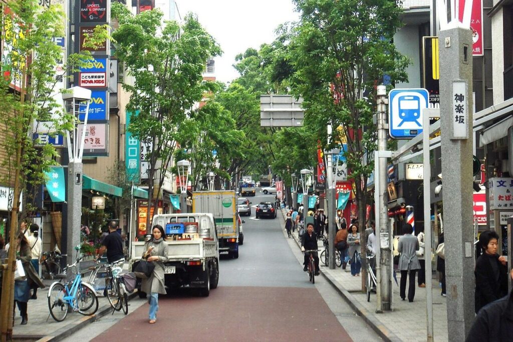 This urban street in Kagurazaka features a mix of vehicle traffic and pedestrians moving along wide sidewalks lined with leafy green trees and numerous commercial signs. Tall, modern light fixtures and a blue train station symbol stand near the foreground, while the road slopes upward toward a densely wooded area in the distance.