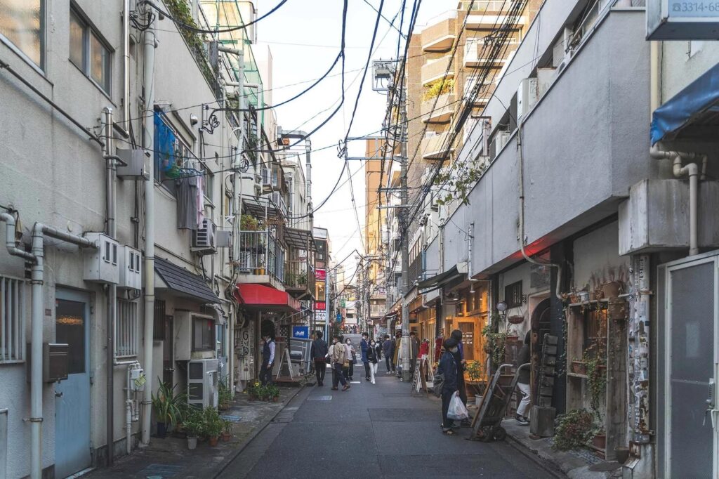 This narrow, urban alleyway in Koenji is lined with weathered multi-story buildings featuring exposed utility pipes, air conditioning units, and small potted plants. A few pedestrians walk along the paved path beneath a dense, chaotic web of overhead power lines that stretch toward the sunlit opening at the end of the street.
