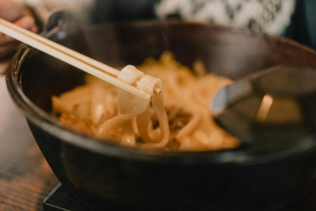 A close-up shot captures a pair of wooden chopsticks lifting thick, flat noodles from a dark bowl, emphasizing the texture of the pasta. The shallow depth of field focuses on the bite in mid-air while softly blurring the savory broth and spoon in the background.