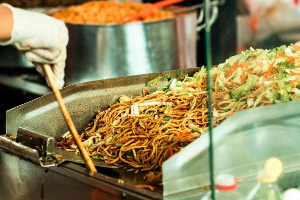A gloved hand uses a long wooden utensil to manage a large quantity of stir-fried noodles mixed with vegetables on a stainless steel flat-top grill. Next to the savory cooked mixture, a fresh mound of chopped cabbage, carrots, and broccoli sits ready for preparation behind a glass partition.